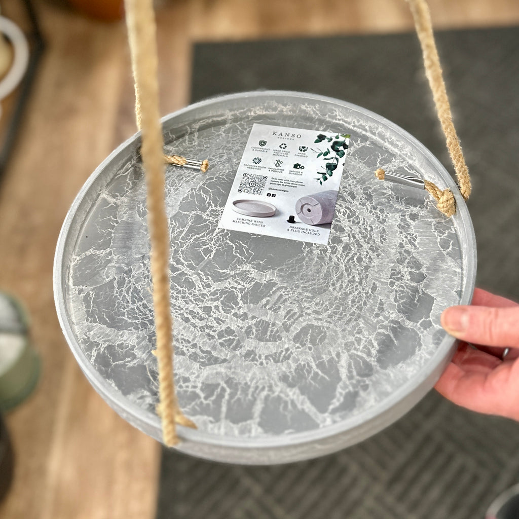 A close-up photo of a hanging gray, marbled plant tray with natural jute rope hangers, held by a hand at Paisley Gardens in Northfield, MN. A product tag for 'KANSO' is attached to the top.