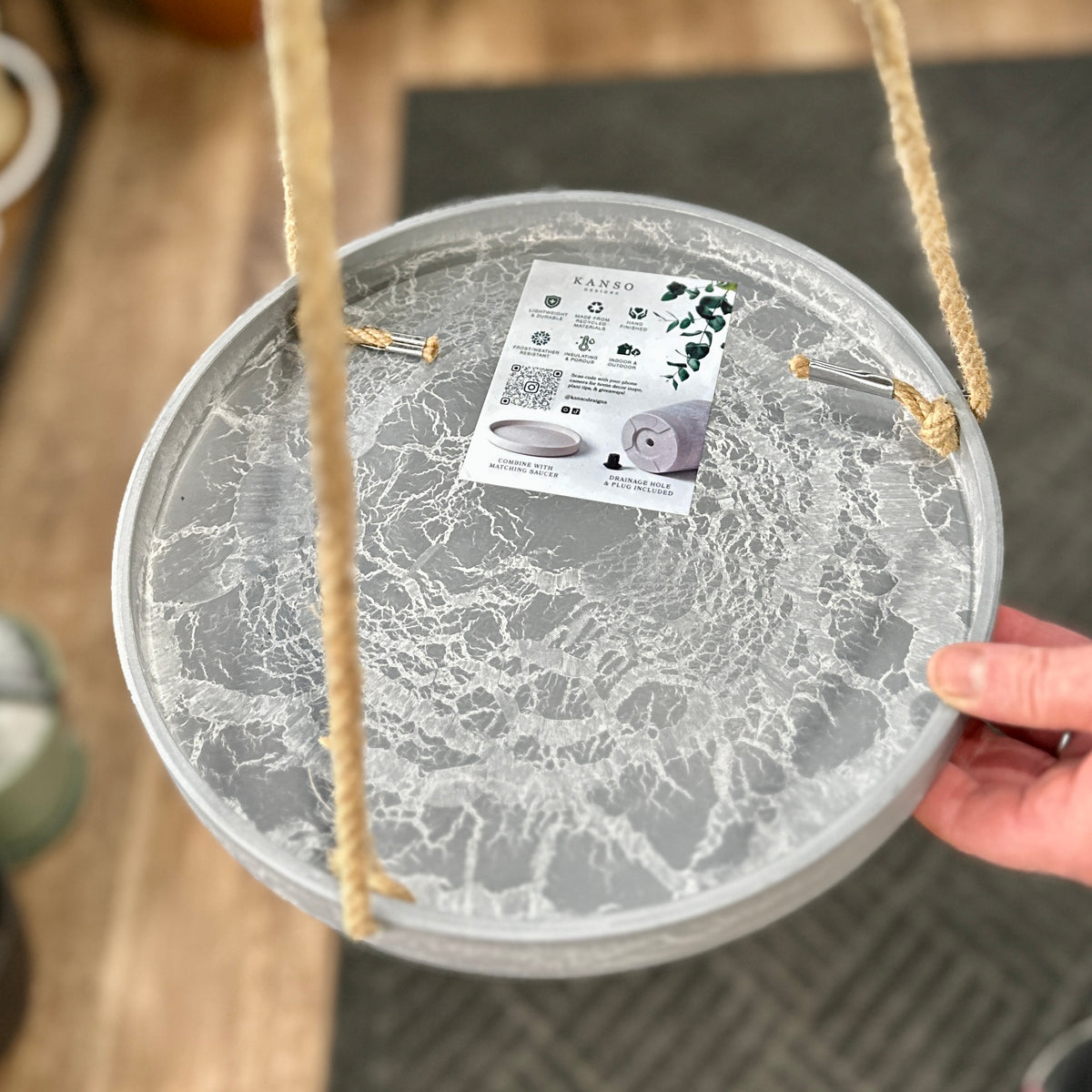 A close-up photo of a hanging gray, marbled plant tray with natural jute rope hangers, held by a hand at Paisley Gardens in Northfield, MN. A product tag for 'KANSO' is attached to the top.