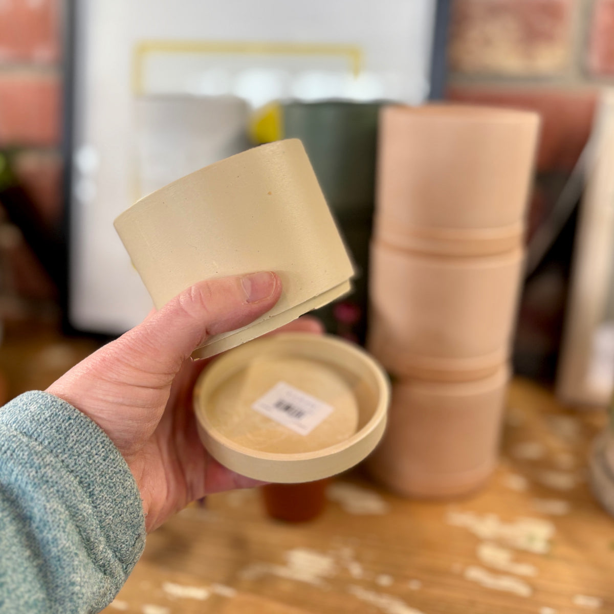 Hand holding a small beige ceramic planter and matching saucer with a blurred background of stacked similar items at Paisley Gardens in Northfield, MN.