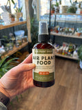 A hand holding a brown spray bottle of air plant food indoors with shelves full of plants at Paisley Gardens in Northfield, MN.
