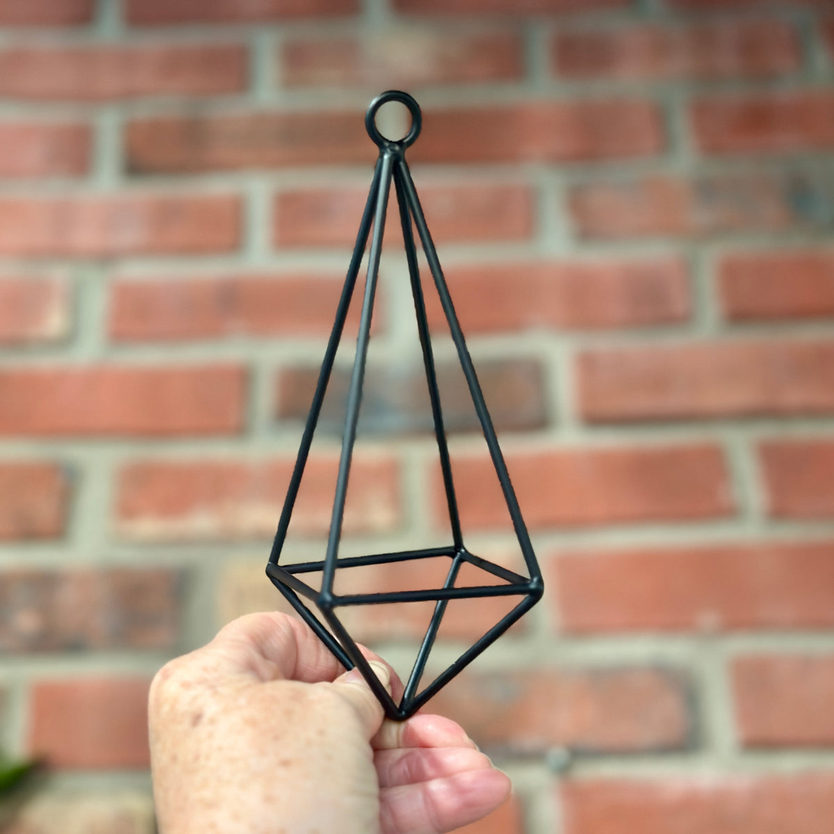 A hand holds a black iron diamond shaped pendant in front of a brick wall at Paisley Gardens in Northfield, MN