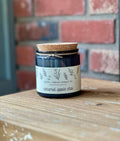 A dark blue glass candle jar with a cork lid and a cream label reading 'Caramel Apple Chai' by Ater + Indigo Candle Co, sitting on a rustic wooden table with a blurred red brick wall in the background at Paisley Gardens in Northfield, MN.