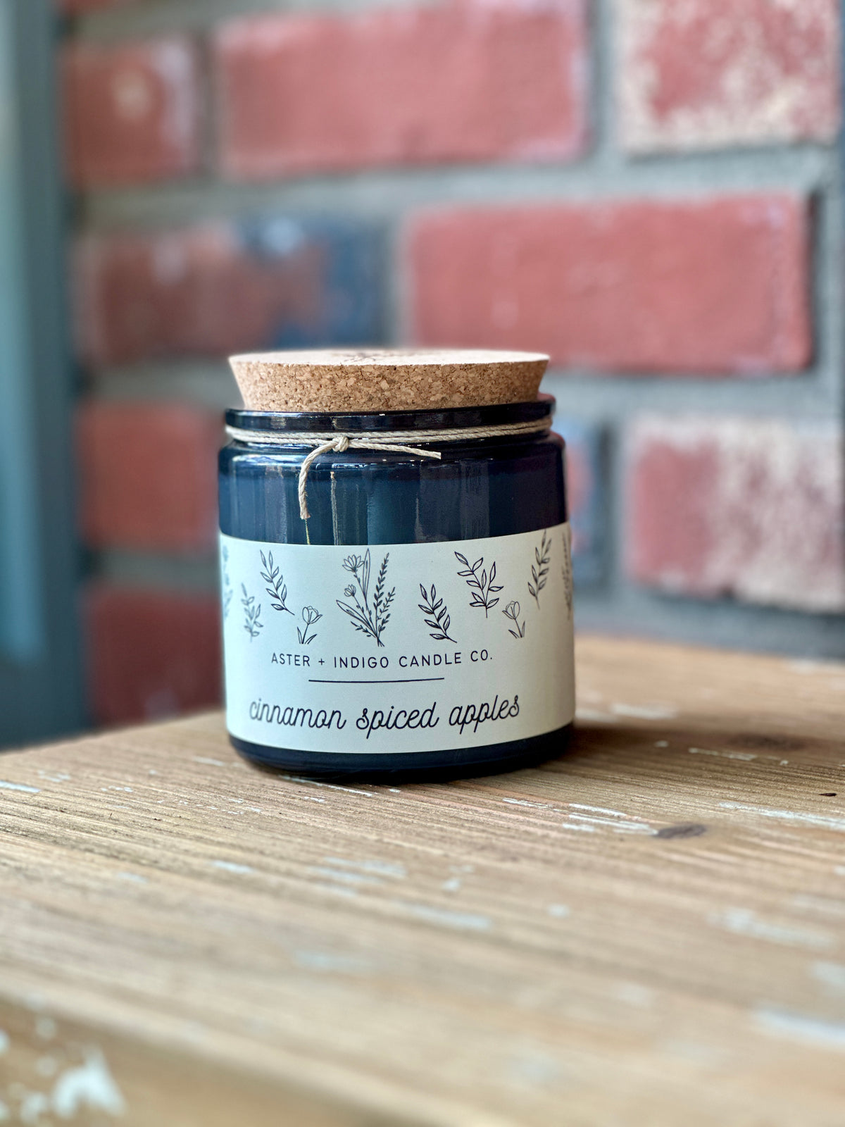 A dark glass jar candle with a cork lid and twine bow sits on a wooden surface in front of a blurred brick wall. The white label reads 'ASTER + INDIGO CANDLE CO. 'Cinnamon Spiced Apples' at Paisley Gardens in Northfield, MN.