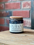 A dark glass jar candle with a cork lid and a white label reading ASTER + INDIGO CANDLE CO. 'Fresh Pressed cCder,' placed on a wooden surface with a blurred red brick wall in the background at Paisley Gardens in Northfield, MN.
