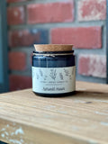 A dark glass jar candle with a cork lid and a cream label reading 'Harvest Moon' and 'ASTER + INDIGO CANDLE CO', placed on a wooden surface with a blurred red brick wall background at Paisley Gardens in Northfield, MN