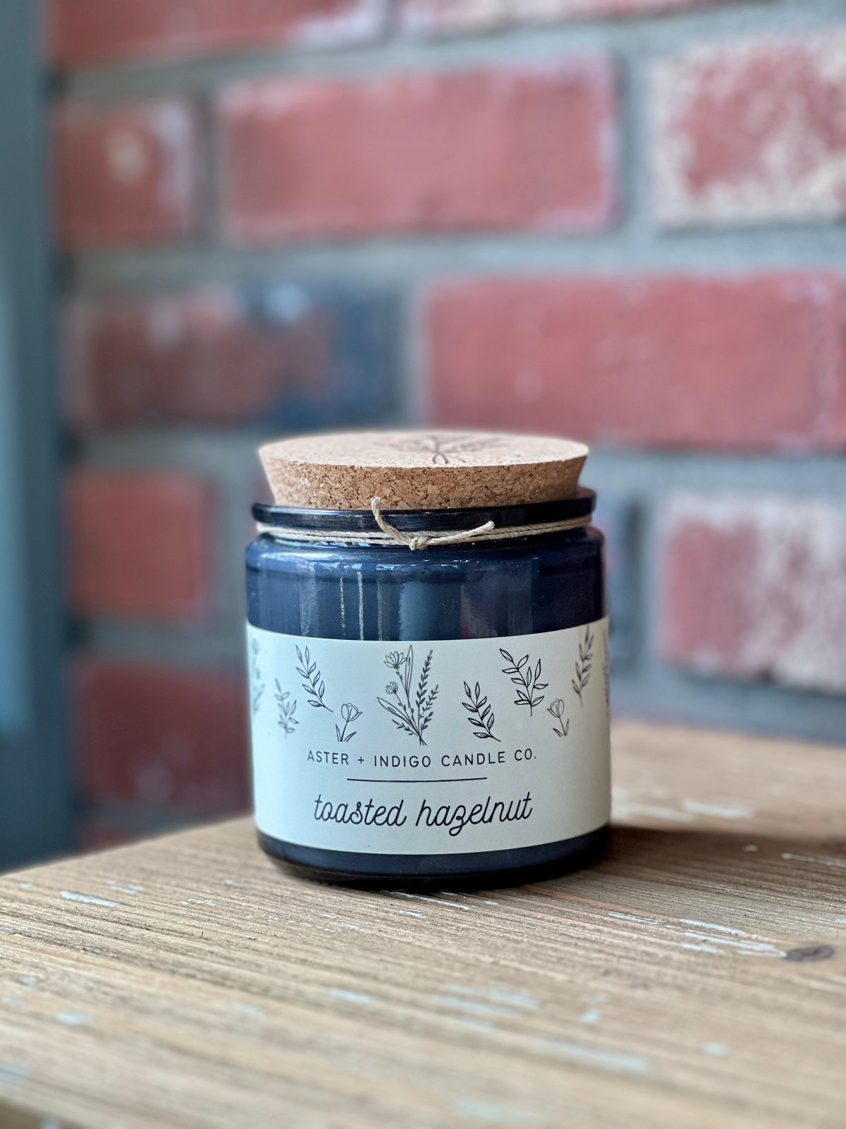 A dark blue glass jar candle with a cork lid and a cream label reading 'Aster + Indigo Candle Co. 'Toasted Hazelnut,' placed on a wooden surface in front of a blurred red brick wall at Paisley Gardens in Northfield, MN.