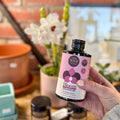 A hand holding a bottle of Bloom liquid fertilizer in front of a blurred white orchid plant at Paisley Gardens in Northfield, MN.