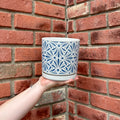 A hand holds a white ceramic planter pot with a blue geometric starburst pattern, set against a red brick wall background at Paisley Gardens in Northfield, MN..