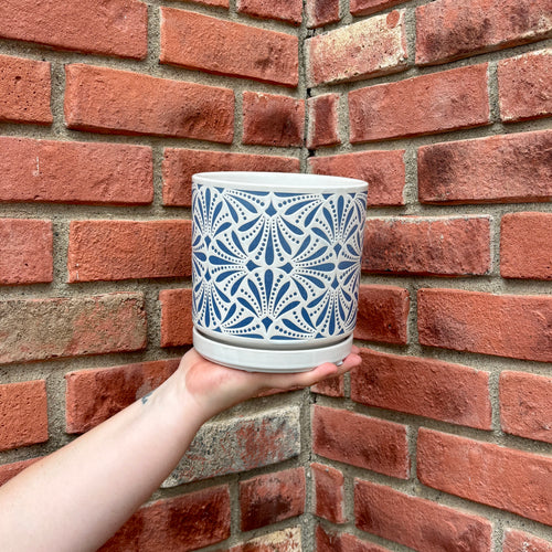 A hand holds a white ceramic planter pot with a blue geometric starburst pattern, set against a red brick wall background at Paisley Gardens in Northfield, MN..