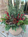 A festive holiday potted arrangement in a large beige pot, featuring green pine and fir branches, natural brown pinecones, and clusters of red and black berries, set outdoors on a stone patio at Paisley Gardens in Northfield, MN.