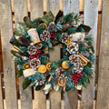 A festive Holiday wreath hanging on a rustic wooden slat door. The wreath is decorated with green and frosted green pine branches, white birch logs, pinecones, cinnamon sticks, dried orange slices, and clusters of red berries at Paisley Gardens in Northfield, MN.