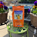 A bag of Coast of Maine Organic & Natural Citrus & Fruit Trees starter fertilizer sits on a green metal plant stand, flanked by potted pansies in an outdoor garden setting at Paisley Gardens in Northfield, MN.