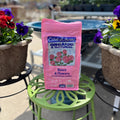 A pink bag of Coast of Maine Organic & Natural Rose & Flower granular fertilizer sits on a green metal plant stand outdoors, flanked by potted pansies at Paisley Gardens in Northfield, MN.