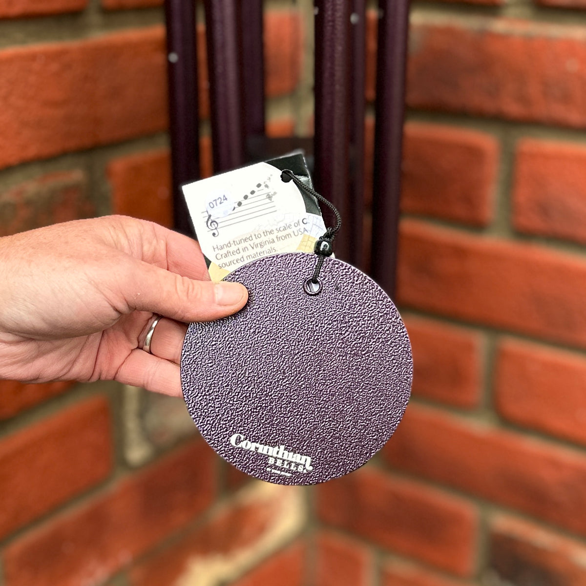 A hanging Corinthian Bells wind chime round textured dark plum wind sail held by a hand in front of a red brick wall at Paisley Gardens in Northfield, MN.