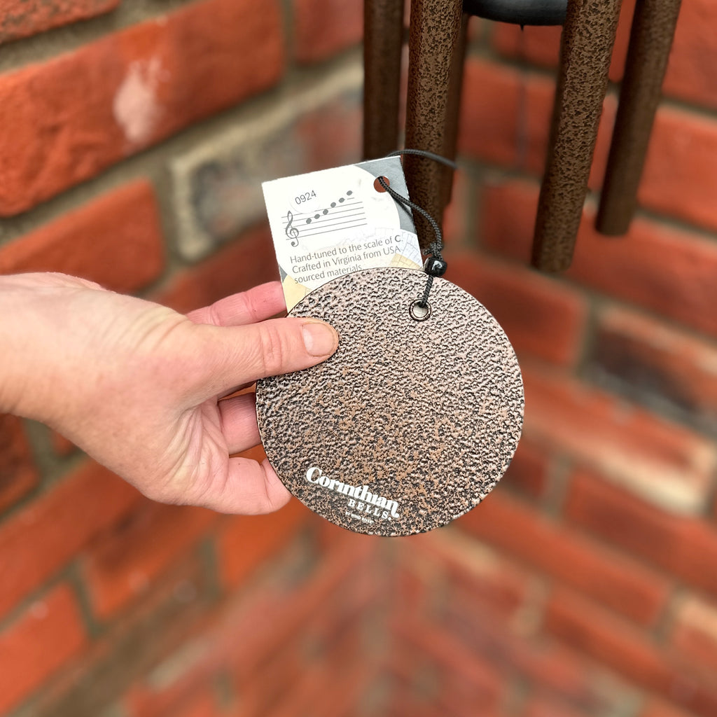 A hand holds a Corinthian Bells round, textured metal wind sail with a hang tag in front of a brick wall aat Paisley Gardens in Northfield, MN.