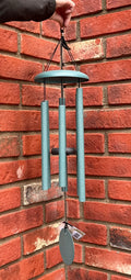 A hanging Corinthian Bells wind chime with Patina Green metal tubes and a matching wind sail,  held by a hand in front of a red brick wall at Paisley Gardens in Northfield, MN.