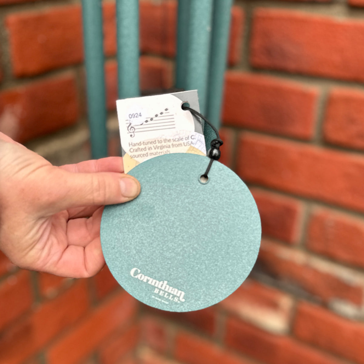 A hand holding a round, textured light blue-green Corinthian Bells wind sail  against a blurred red brick wall background at Paisley Gardens in Northfield, MN.