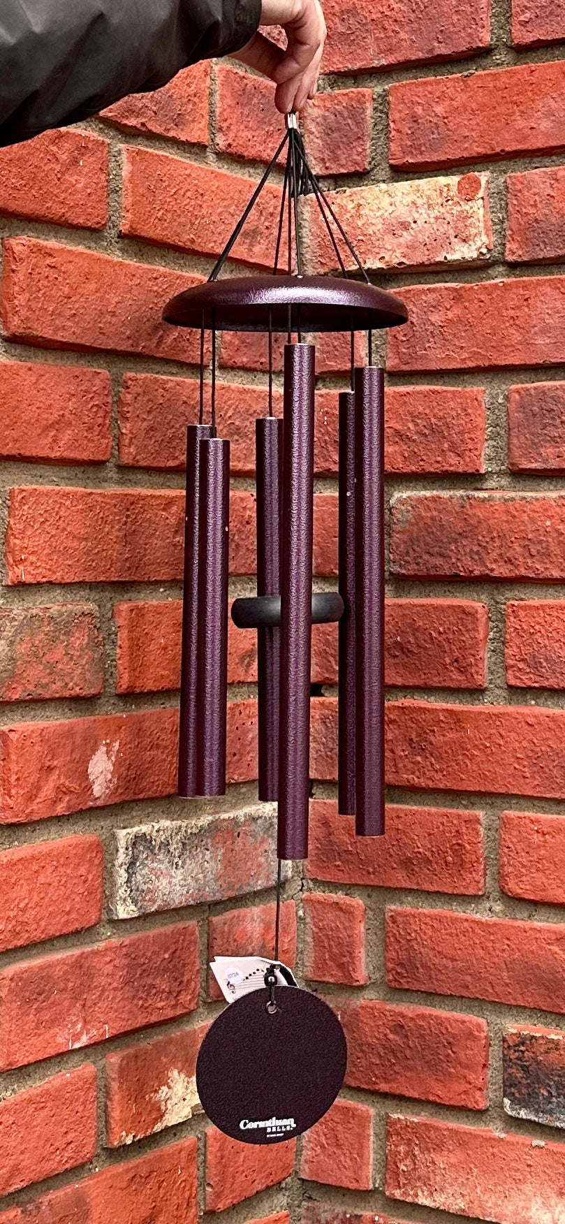 A hanging Corinthian Bells wind chime with dark plum metal tubes and a matching wind sail,  held by a hand in front of a red brick wall at Paisley Gardens in Northfield, MN.