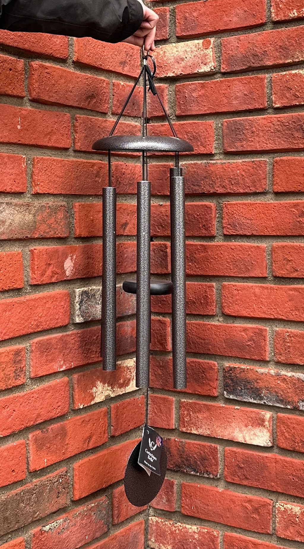 A hanging Corinthian Bells wind chime with dark silver gray metal tubes and a matching wind sail,  held by a hand in front of a red brick wall at Paisley Gardens in Northfield, MN.