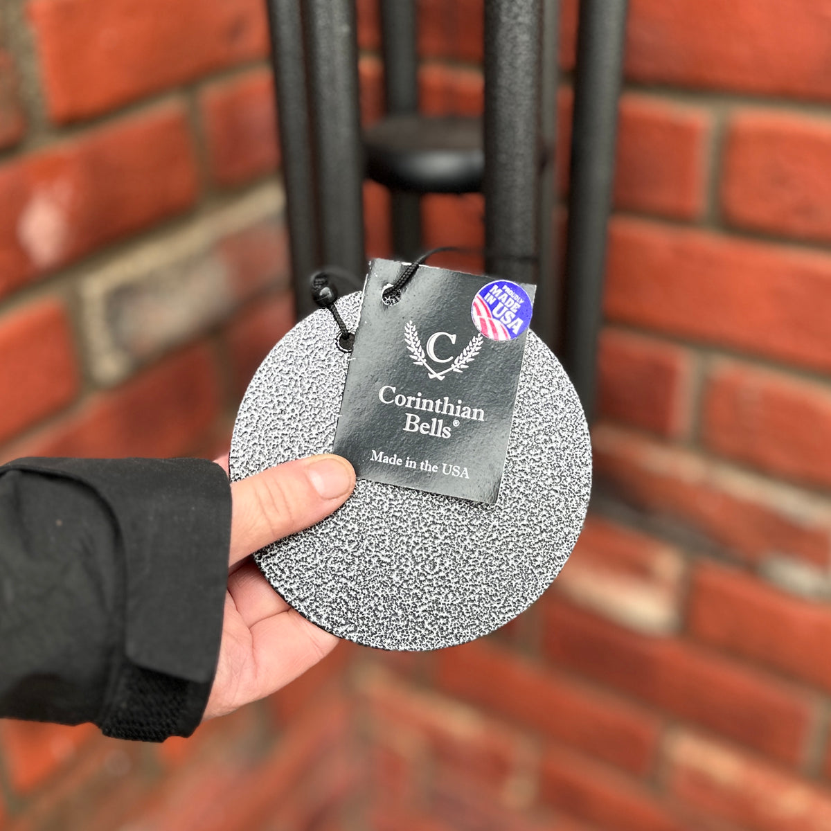 A hanging Corinthian Bells dark gray, round textured wind sale with a black tag held by a hand in front of a red brick wall at Paisley Gardens in Northfield, MN.