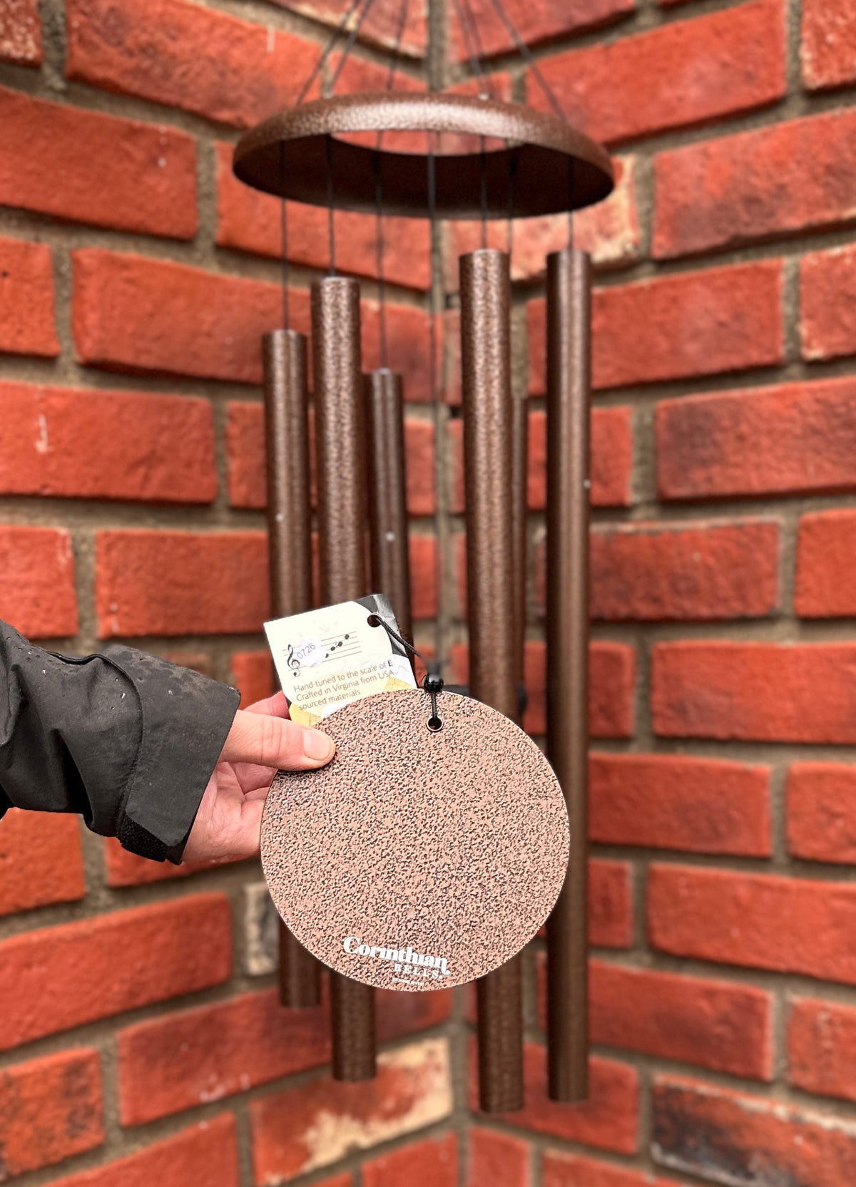 A copper wind chime with a circular wind sail, printed with 'Corinthian Bells' shown against a red brick wall at Paisley Gardens in Northfield, MN.