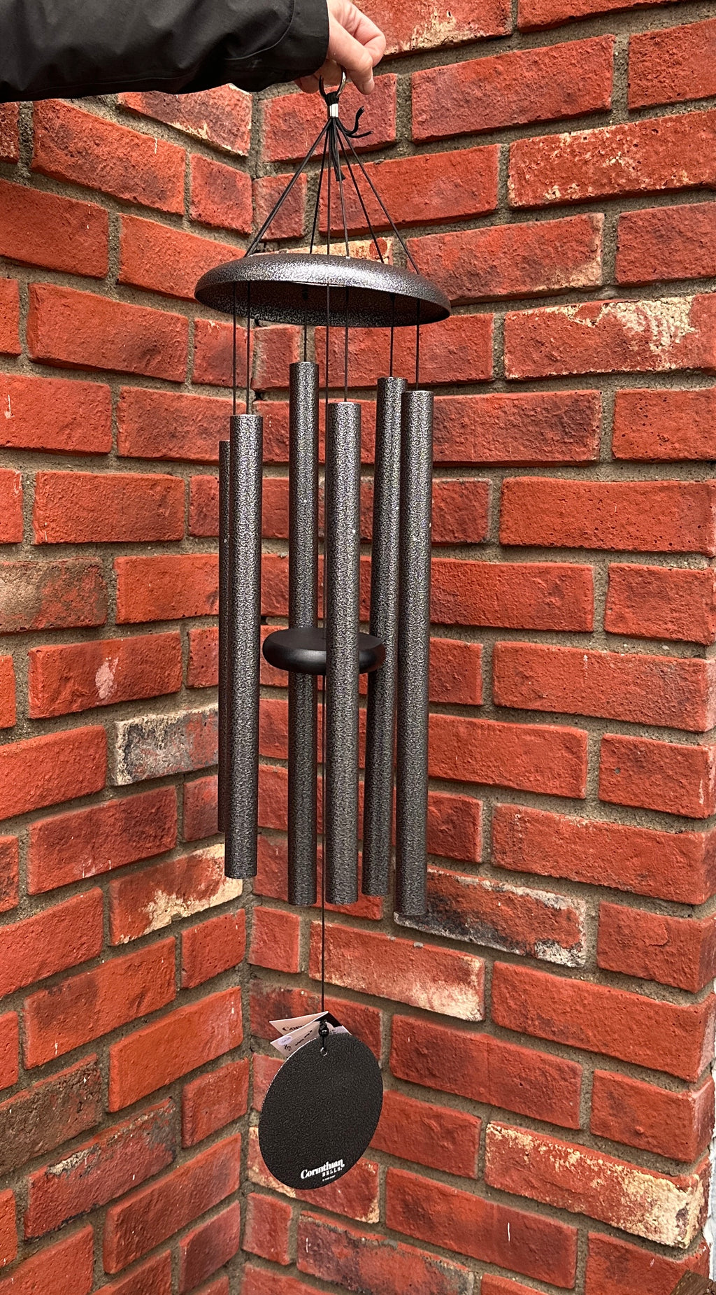 A hanging Corinthian Bells wind chime with silver gray metal tubes and a matching wind sail, held by a hand in front of a red brick wall at Paisley Gardens in Northfield, MN.