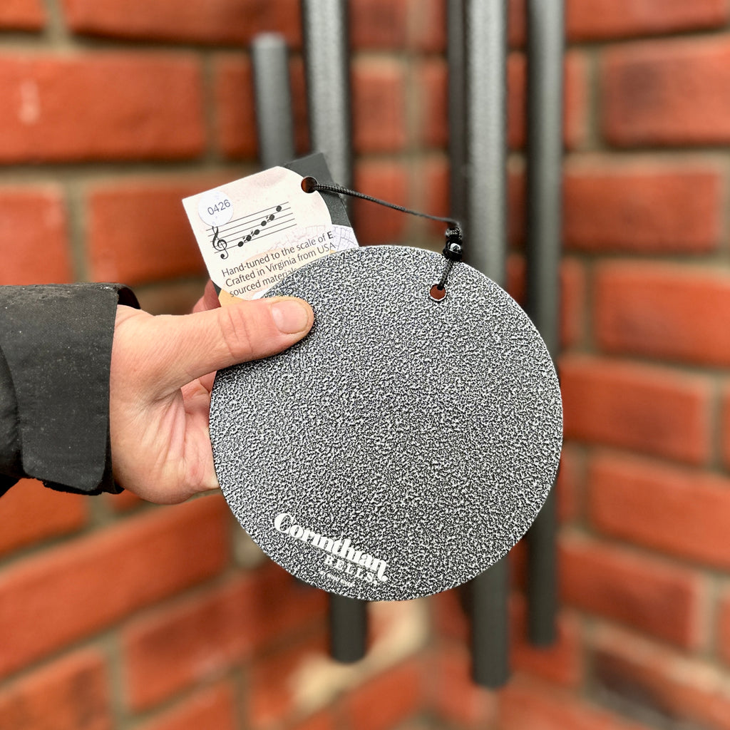 A hanging Corinthian Bells silver gray, round textured wind sail and tag,  held by a hand in front of a red brick wall at Paisley Gardens in Northfield, MN.