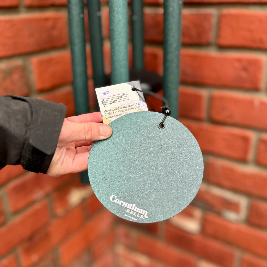 A hand holding a round, textured green  wind sail, for a wind chime, with the brand name 'Corinthian Bells' printed on it, set against a blurred brick wall background at Paisley Gardens, in Northfield, MN.