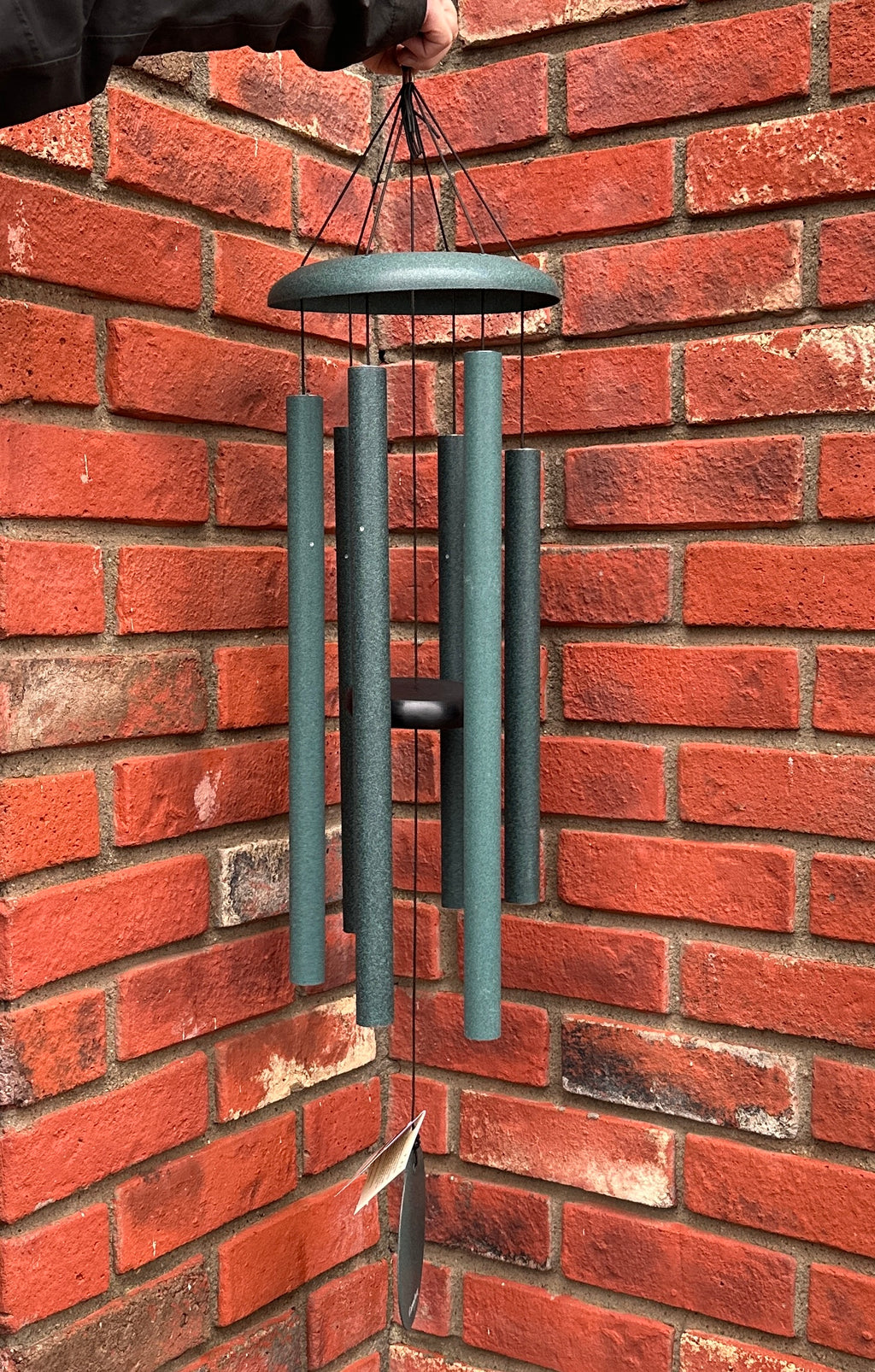 A hand is holding a dark green metal wind chime with multiple hanging tubes and a striker bar, suspended against a red brick wall at Paisley Gardens in Northfield, MN.