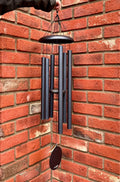 A deep blue metal wind chime with six vertical tubes and a hanging striker, held by a hand against a red brick wall corner at Paisley Gardens in Northfield, MN.