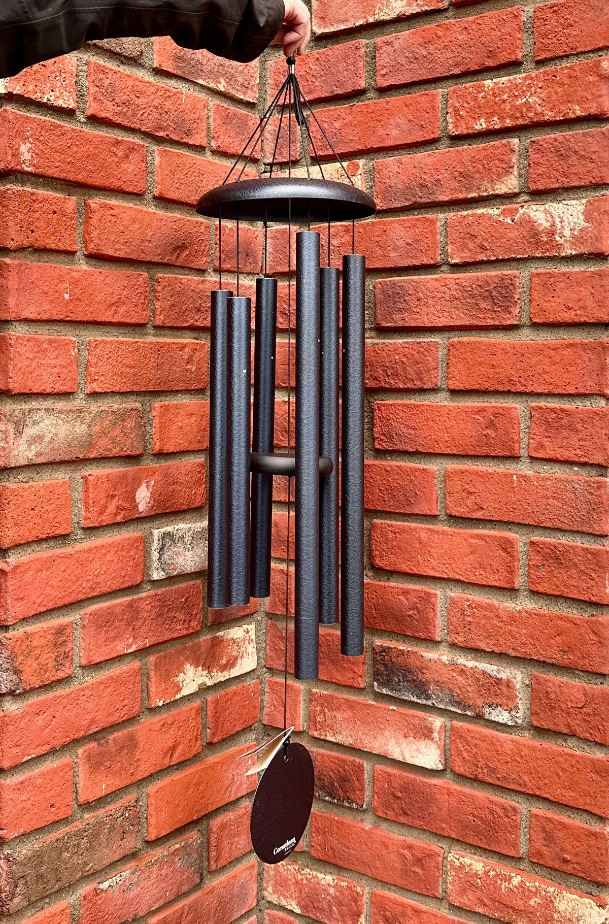 A deep blue metal wind chime with six vertical tubes and a hanging striker, held by a hand against a red brick wall corner at Paisley Gardens in Northfield, MN.