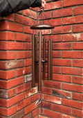 A person's hand holds a metal wind chime with dark bronze-colored tubes and a matching top cup, hanging against a red brick wall corner at Paisley Gardens in Northfield, MN.