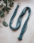 Deep green macrame plant hanger on a light wooden surface with green leaves at Paisley Gardens in Northfield, MN.