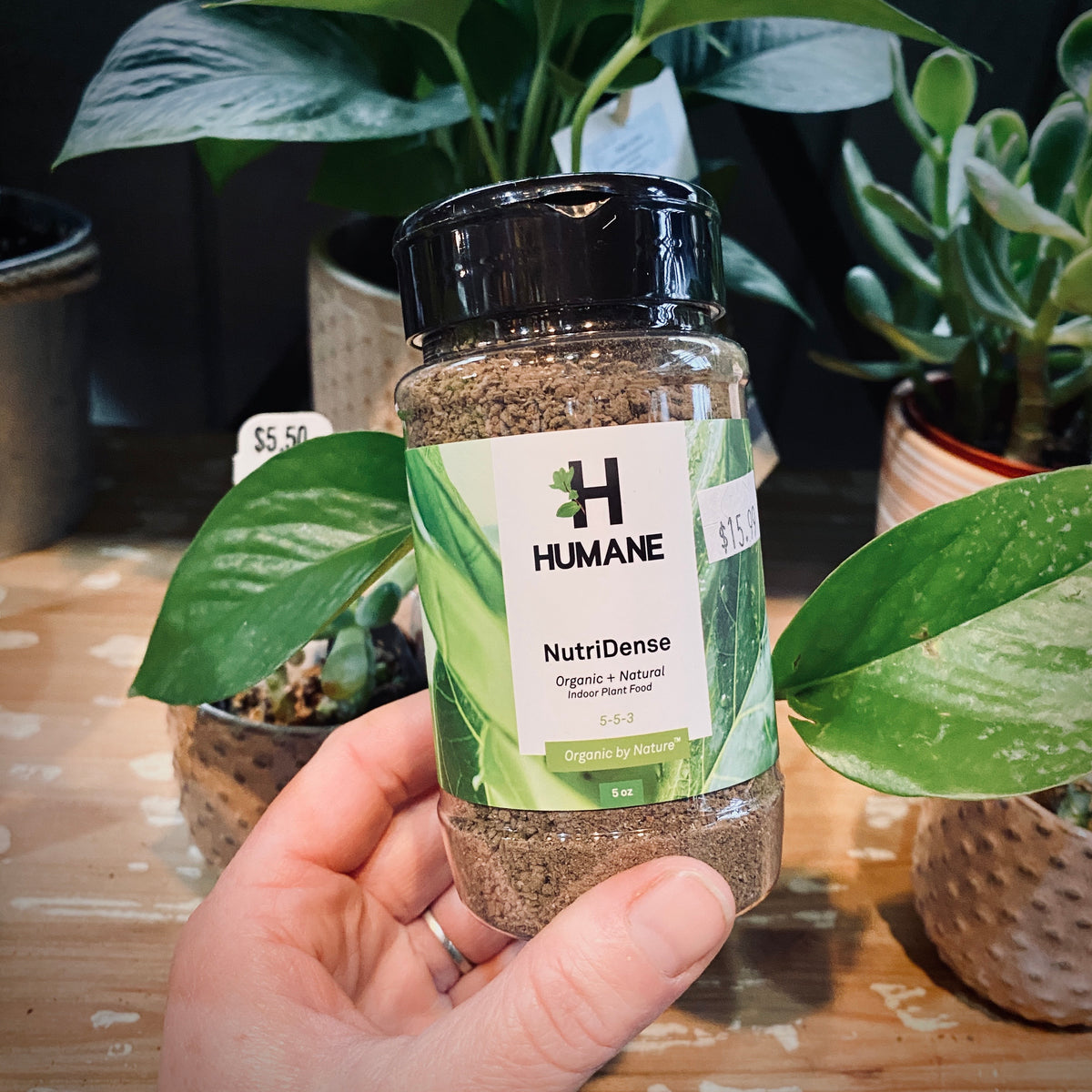 A hand holds a bottle of Humane NutriDense organic and natural indoor fertilizer in front of a background of potted green plants at Paisley Gardens in Northfield, MN.