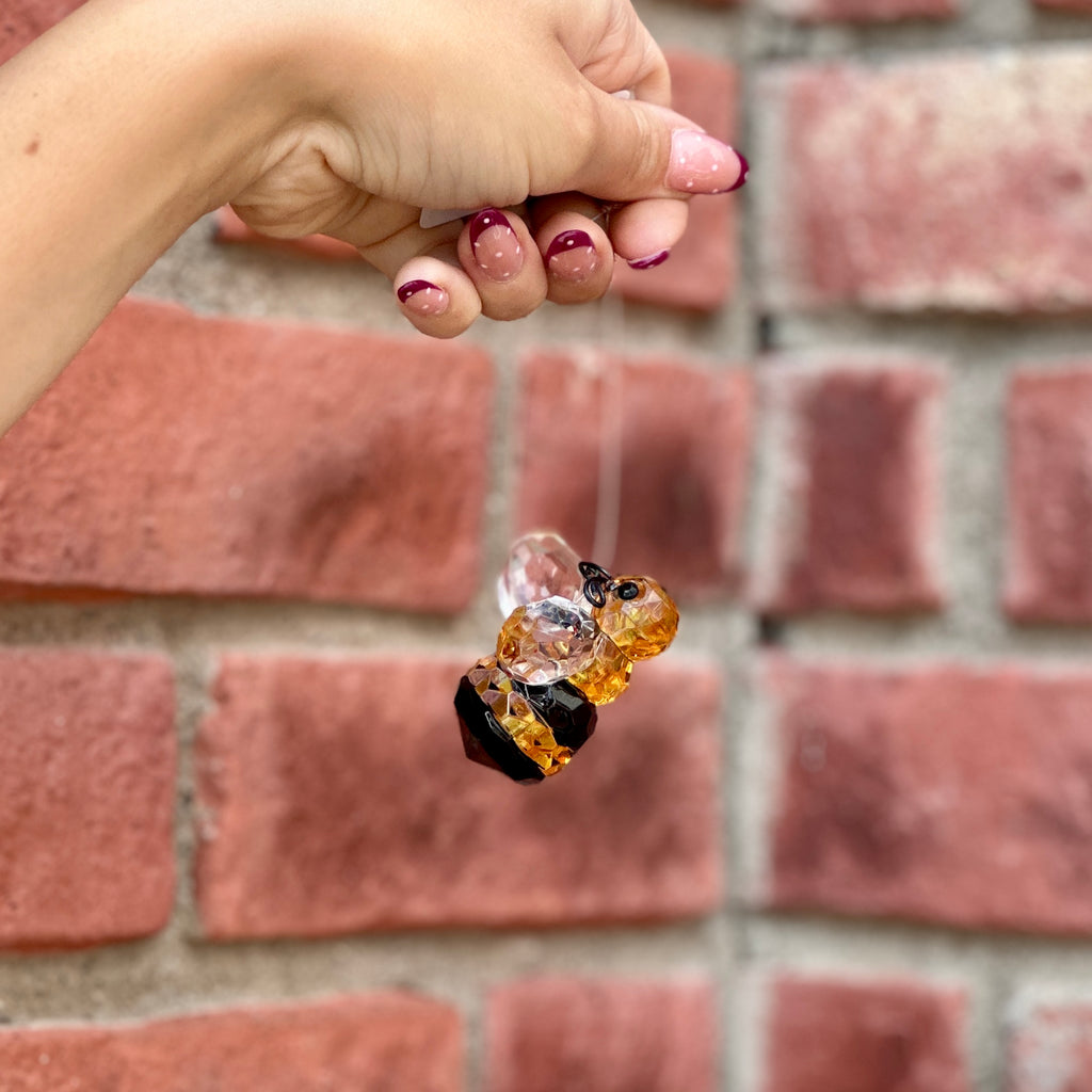 A manicured hand holds a small hanging  honey bee ornament made from amber yellow and  black faceted acrylic in front of a blurred red brick wall at Paisley Gardens in Northfield, MN.
