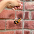 A manicured hand holds a small hanging  honey bee ornament made from amber yellow and  black faceted acrylic in front of a blurred red brick wall at Paisley Gardens in Northfield, MN.