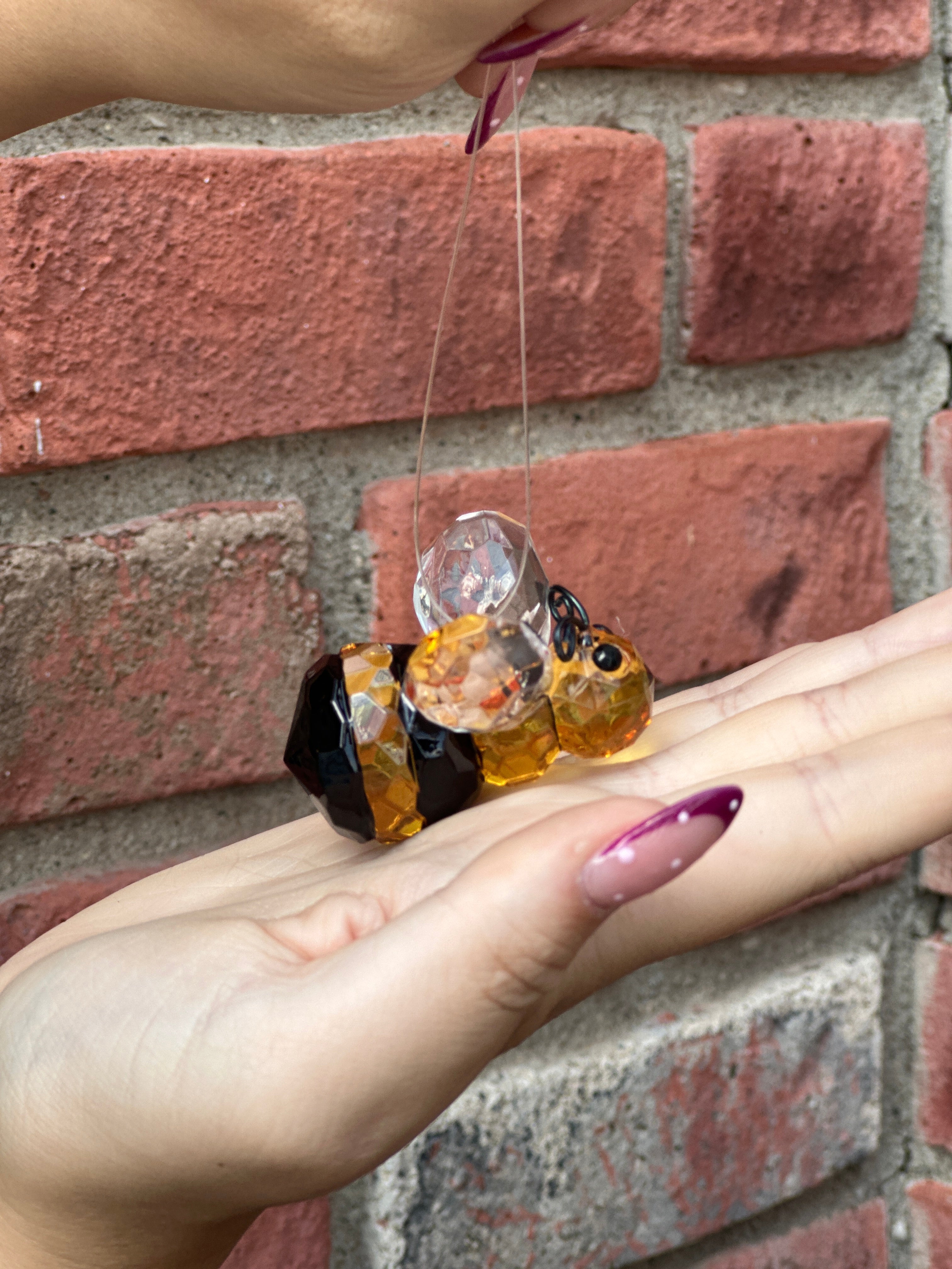 A hand holding a small acrylic bee-shaped ornament with amber yellow, black, and clear faceted details, positioned against a red brick wall background at Paisley Gardens in Northfield, MN.