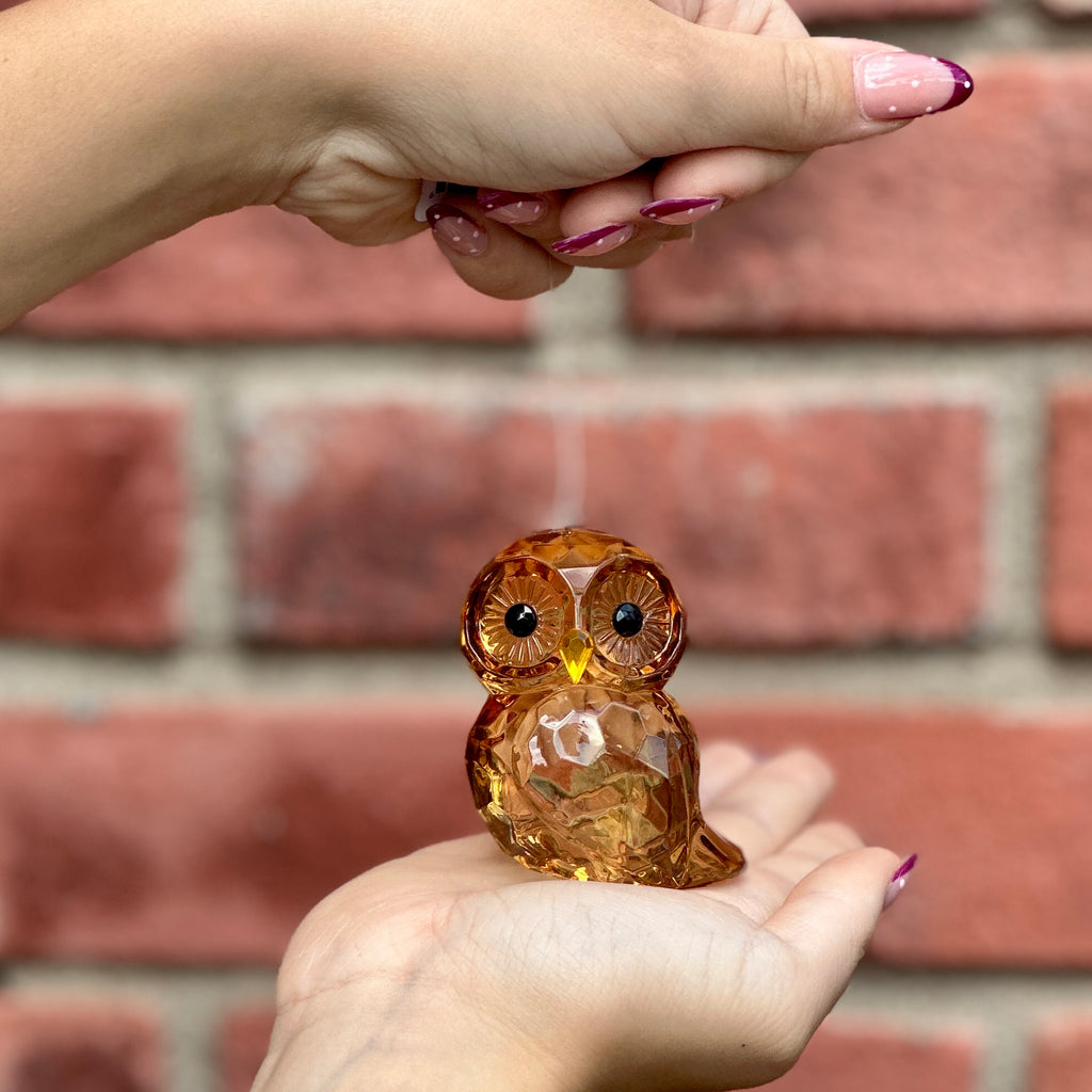 A small faceted amber owl figurine being held in a person's hands against a blurred red brick wall background at Paisley Gardens in Northfield, MN.