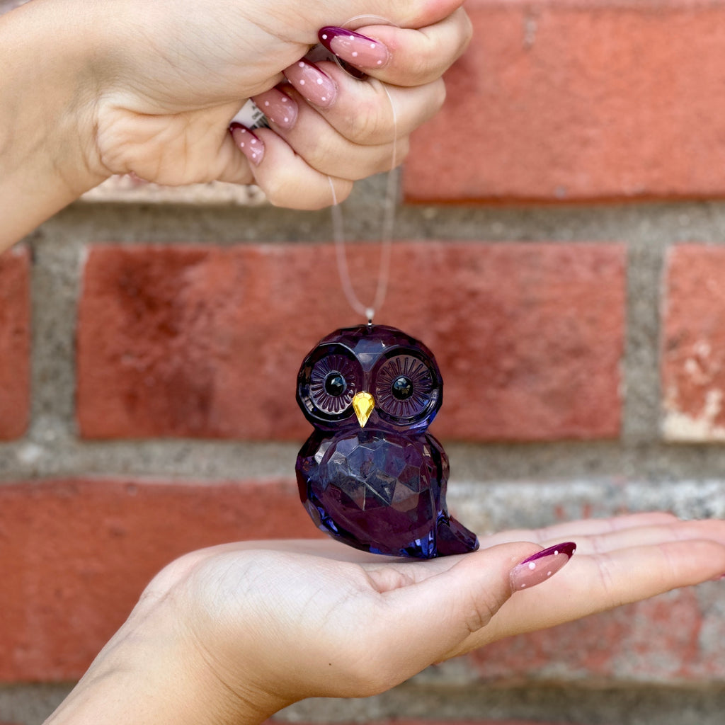 A faceted blue owl ornament being held in a person's hands against a red brick wall background at Paisley Gardens in Northfield, MN.