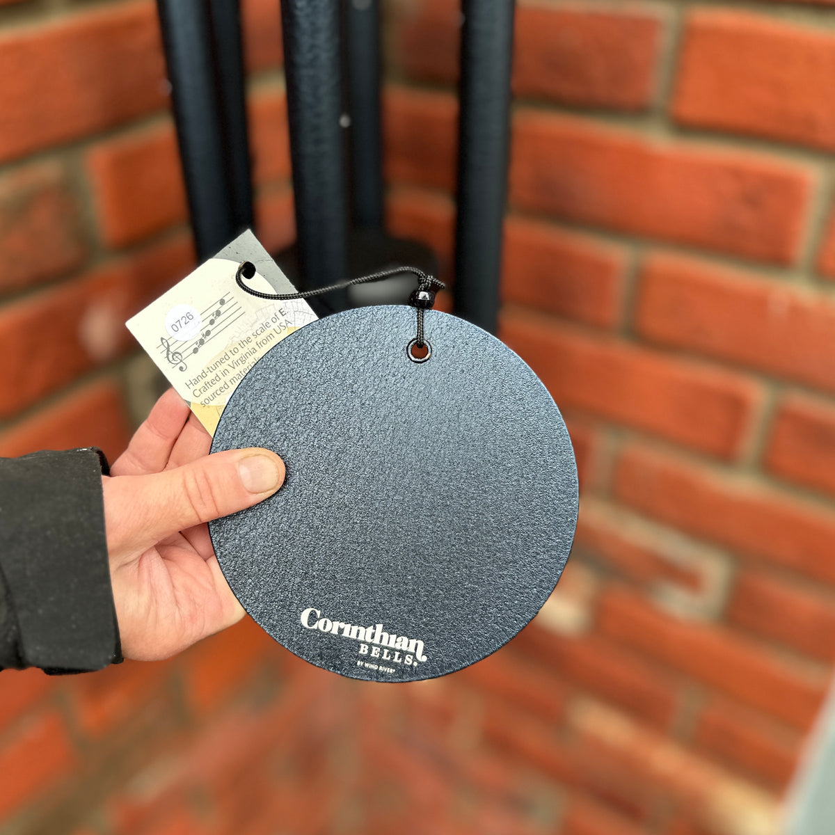 A hand holding a round, textured dark blue wind sail, for a wind chime, with the brand name 'Corinthian Bells' printed on it, set against a blurred brick wall background at Paisley Gardens, in Northfield, MN.