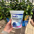 A hand holds a white plastic tub of Jack's Classic Hydrangea Blue water soluble plant food against a background of green hydrangea bushes outdoors.