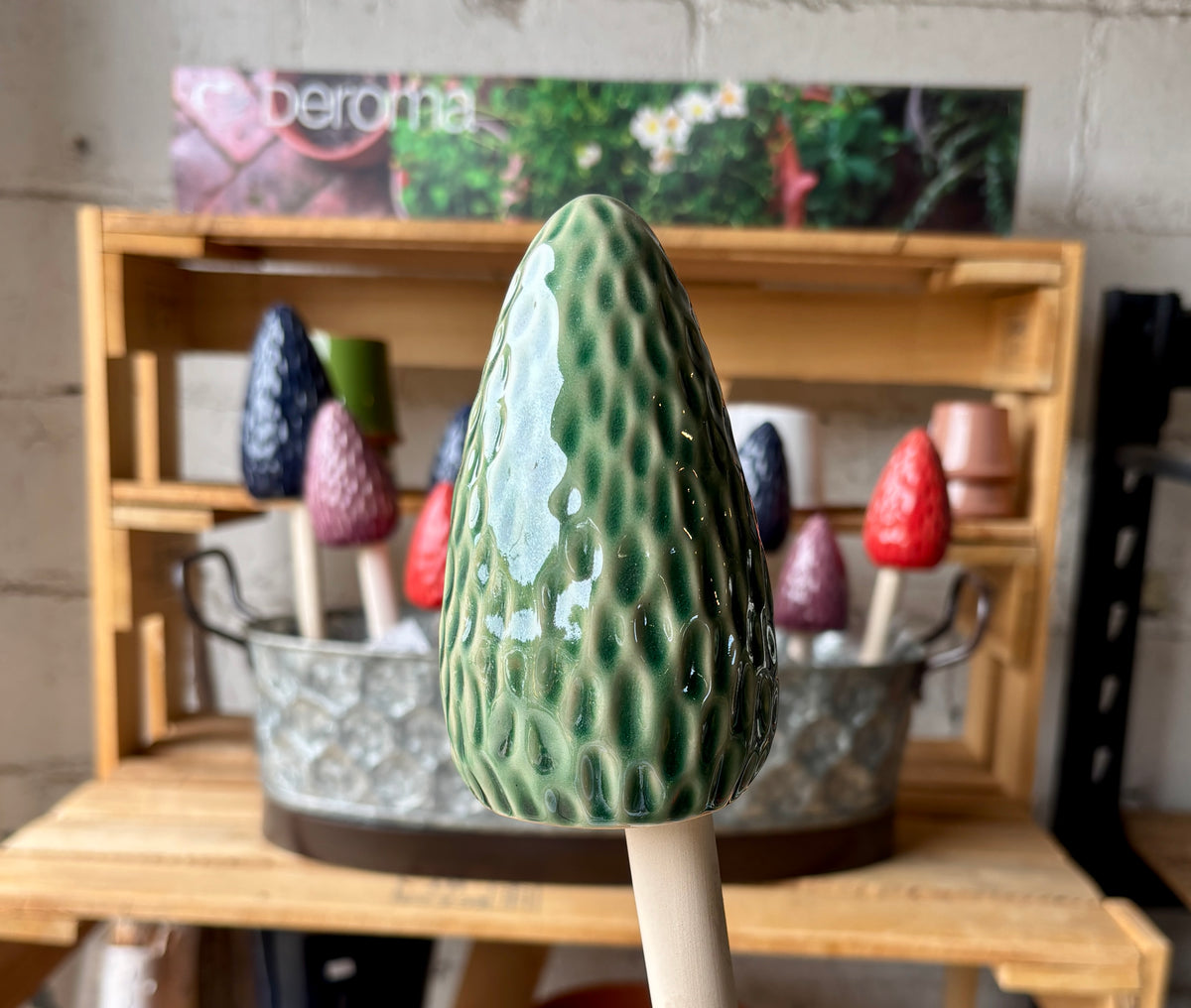 A green glazed ceramic mushroom topper with a textured, wavy surface and a light-colored wooden handle, shown in a workshop setting with other colorful ceramic mushrooms in the background. at Paisley Gardens in Northfield, MN.