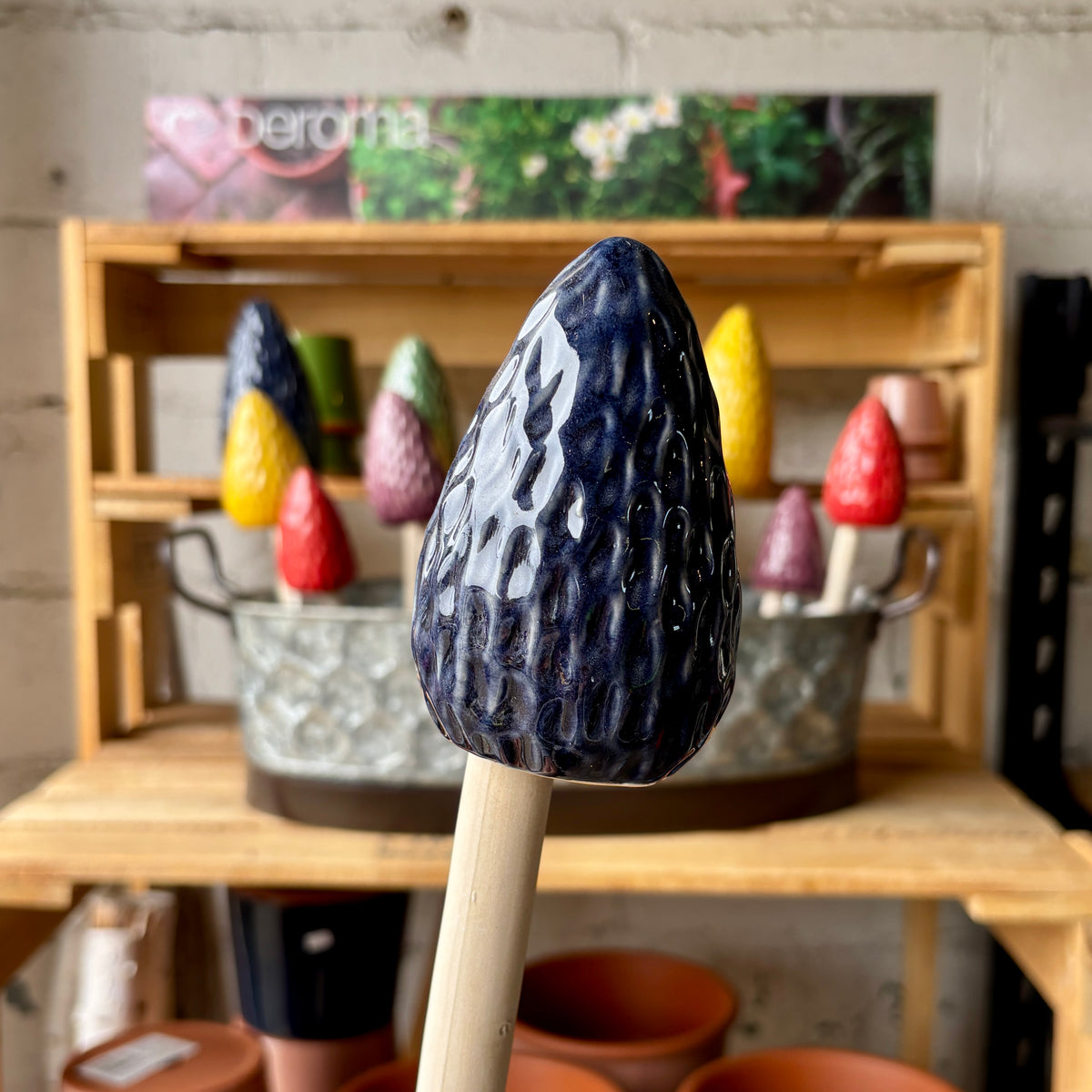 A close-up of a dark navy blue, morel mushroom garden stake with a textured, glossy surface, set against a blurred background of a wooden shelf holding other colorful morel mushroom stakes at Paisley Gardens in Northfield, MN..
