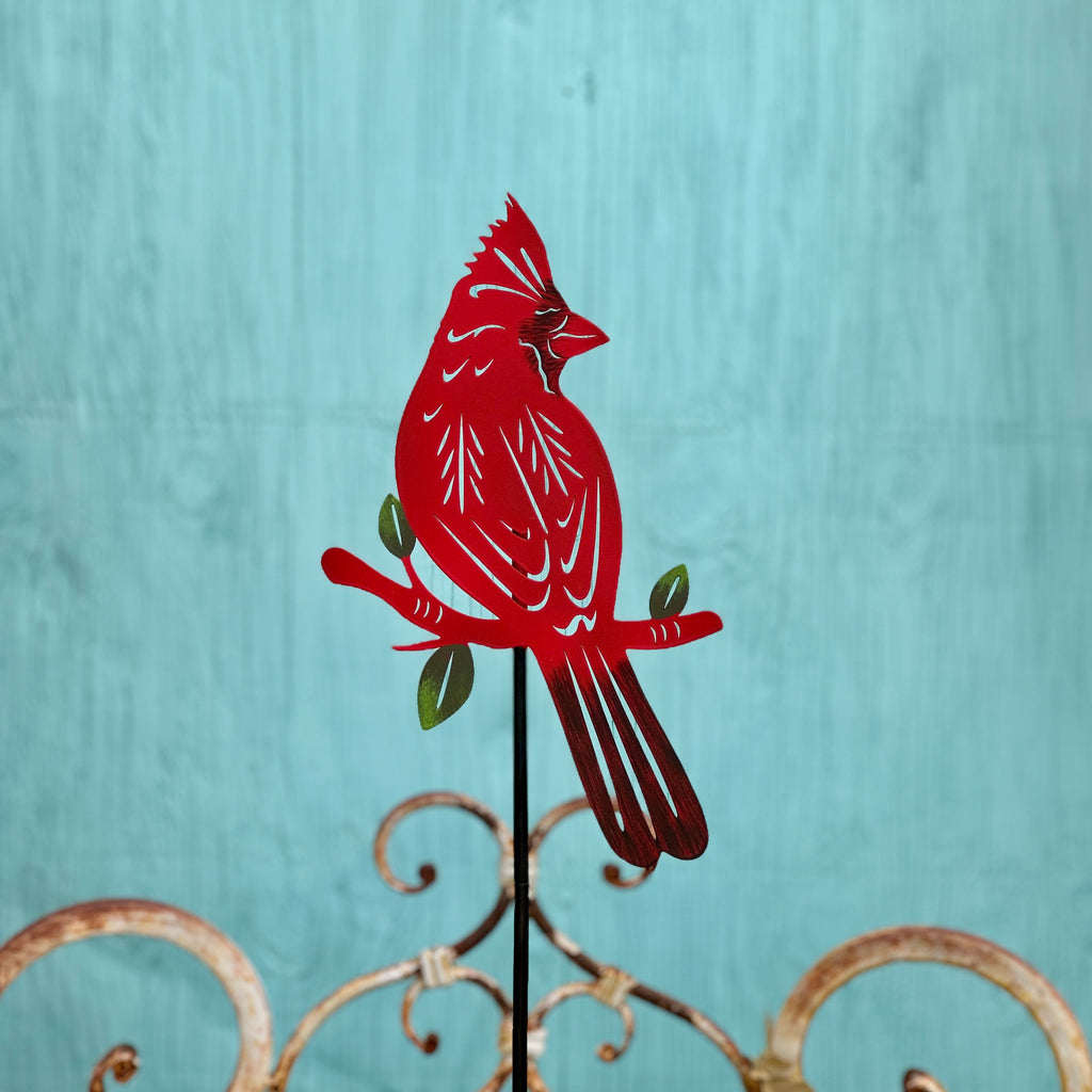 A red metal cardinal garden stake with laser cut details,  perched on a branch with green leaves, set against a textured turquoise background at Paisley Gardens in Northfield, MN.