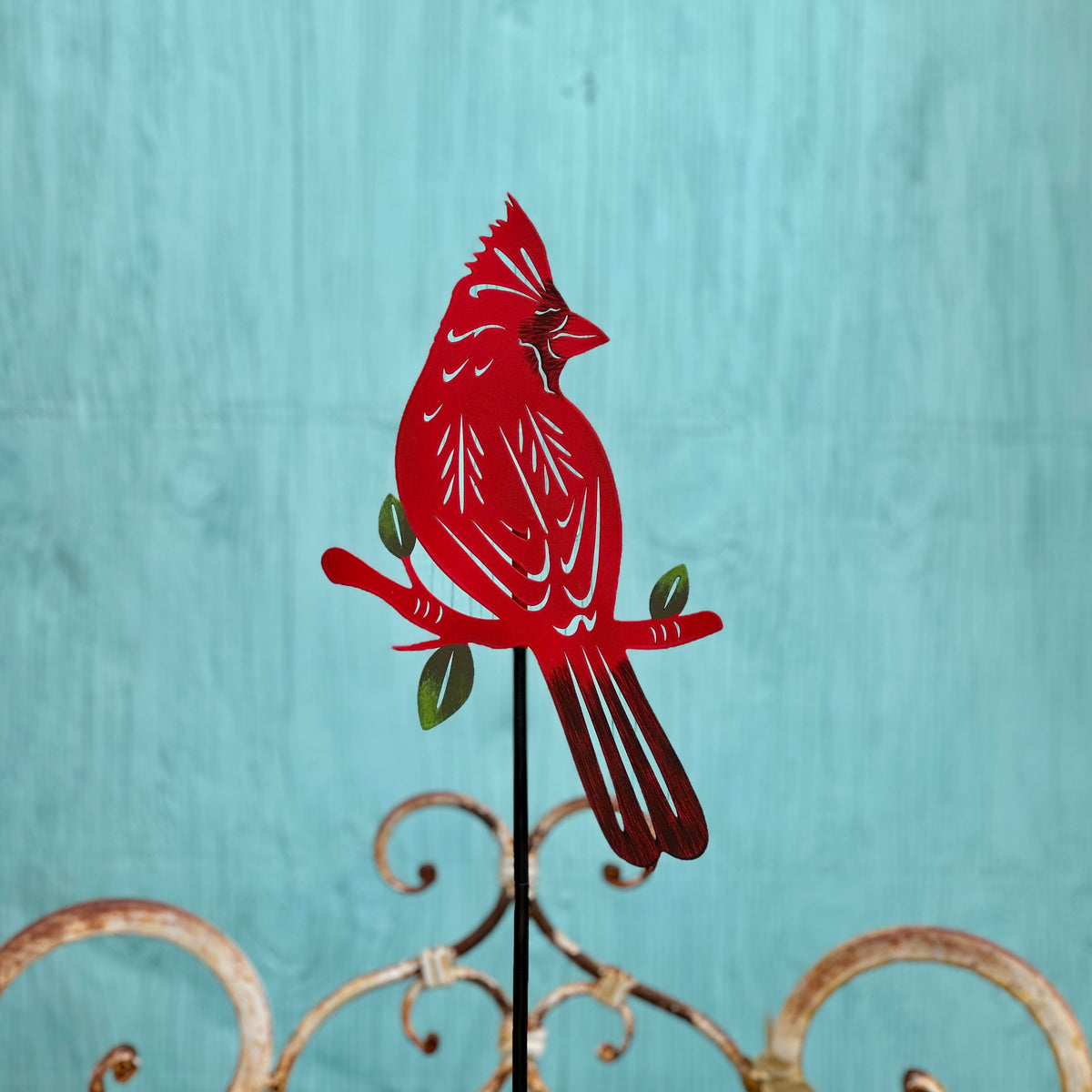 A red metal cardinal garden stake with laser cut details,  perched on a branch with green leaves, set against a textured turquoise background at Paisley Gardens in Northfield, MN.
