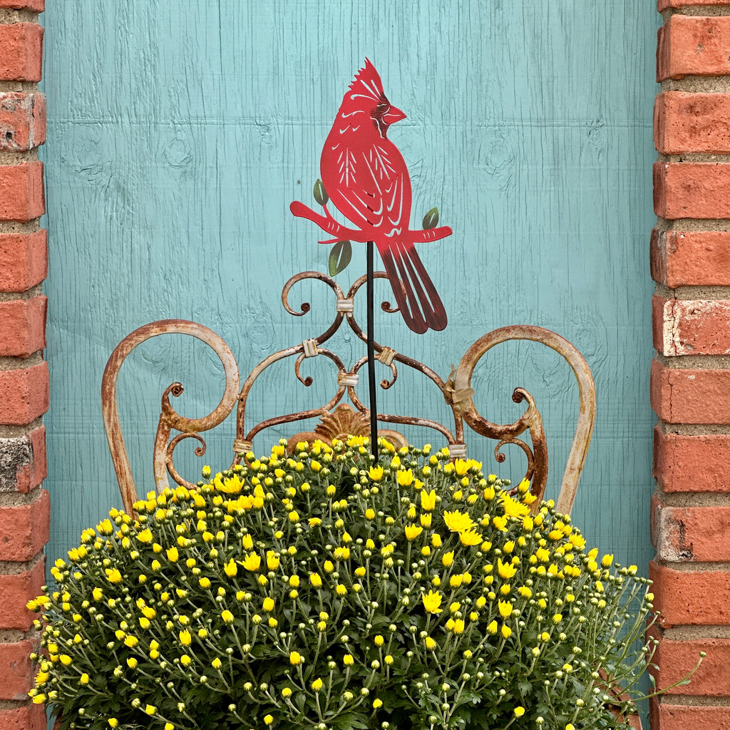 A red cardinal metal garden stake with green leaves, displayed in a large yellow mums flower bed in front of a rustic turquoise wooden door framed by red brick at Paisley Garden in Northfield, MN.