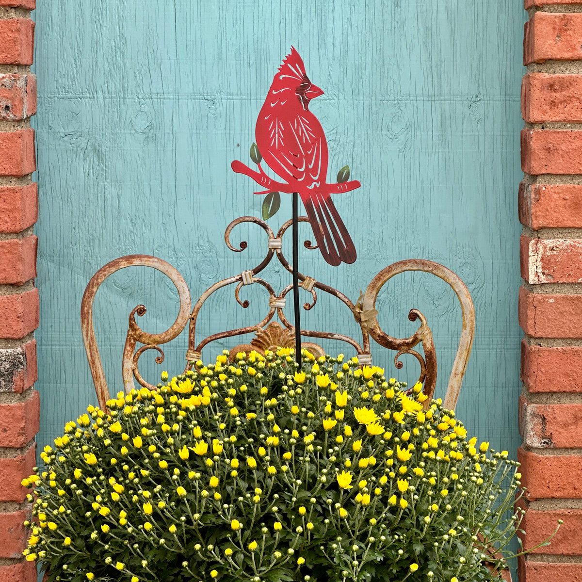 A red cardinal metal garden stake with green leaves, displayed in a large yellow mums flower bed in front of a rustic turquoise wooden door framed by red brick at Paisley Garden in Northfield, MN.