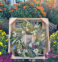 A dried flower wreath with sage green, white, purple, and pink botanicals, displayed inside a light wood crate in a garden center surrounded by colorful flowers at Paisley Gardens in Northfield, MN.