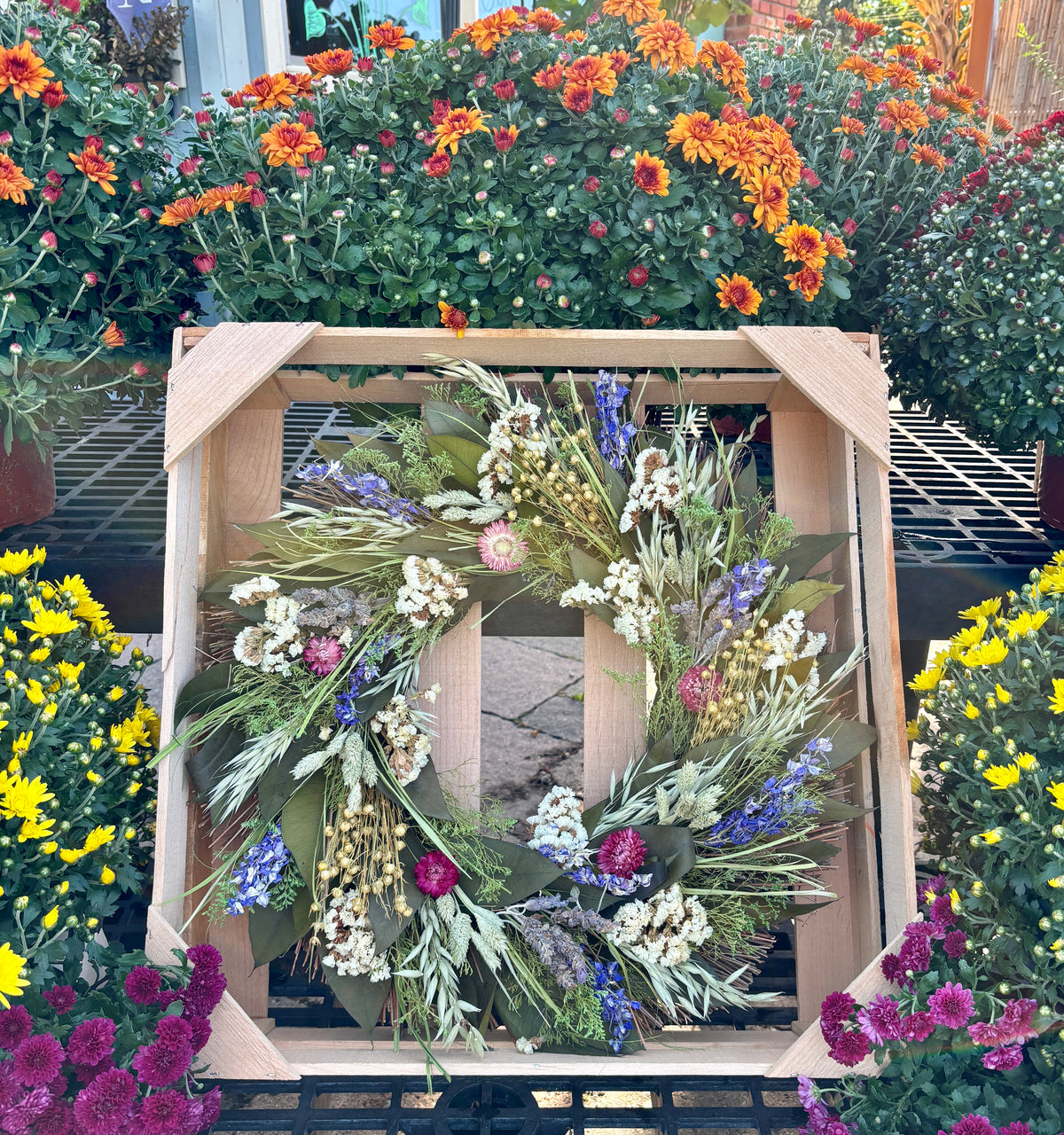 A dried flower wreath with sage green, white, purple, and pink botanicals, displayed inside a light wood crate in a garden center surrounded by colorful flowers at Paisley Gardens in Northfield, MN.