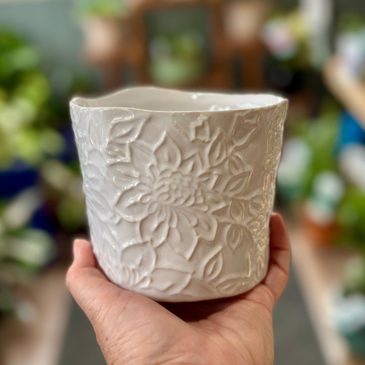 A hand holding a small, white ceramic planter with embossed floral patterns, set against a blurred background of green plantsat Paisley Gardens in Northfield, MN.